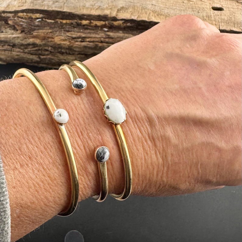 May include: Gold-toned metal bracelets with white and gray stone accents. The bracelets have a simple, elegant design, with the stones set in a minimalist style. The bracelets are shown on a person's wrist.