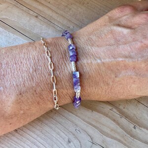 Puede incluir: Una pulsera con cuentas c&uacute;bicas de amatista morada intercaladas con peque&ntilde;os espaciadores de metal plateado, usada junto a una pulsera de cadena delicada de color plateado con eslabones ovalados alargados en una mu&ntilde;eca.