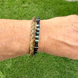 May include: Two bracelets on a wrist. One is a braided leather bracelet in shades of brown and tan. The other is a beaded bracelet with black beads and turquoise-colored stones. The bracelets are worn against a background of green grass.