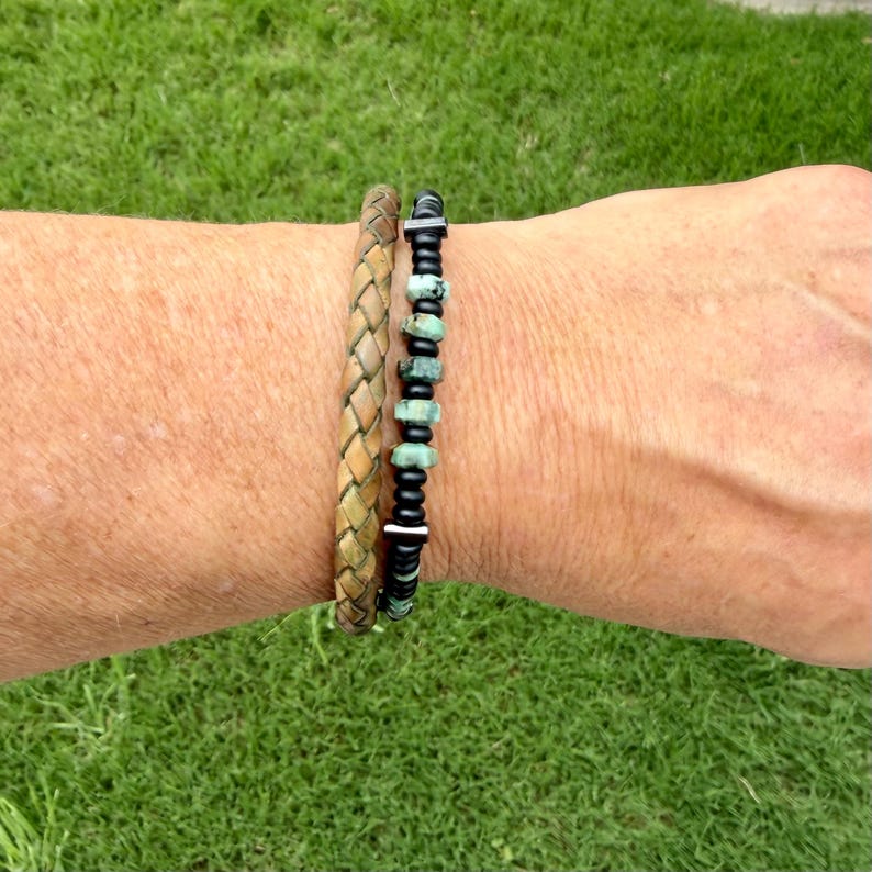 May include: Two bracelets on a wrist. One is a braided leather bracelet in shades of brown and tan. The other is a beaded bracelet with black beads and turquoise-colored stones. The bracelets are worn against a background of green grass.