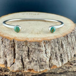 K&ouml;nnte beinhalten: Ein silberfarbenes Armreif-Armband mit t&uuml;rkisfarbenen Steinen an jedem Ende. Das Armband wird auf einer rustikalen Holzoberfl&auml;che pr&auml;sentiert, die den Kontrast von Metall und Edelsteinen hervorhebt. Ein leicht zu tragendes, stilvolles Accessoire.