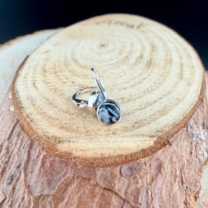 May include: A pair of silver leverback earrings with round, black and white patterned stones. The earrings are displayed on a wooden surface with a natural wood grain pattern.
