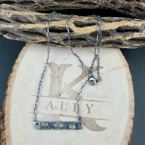 May include: A silver-toned metal necklace featuring a rectangular bar pendant with a textured surface and three small, round and square green stones. The necklace has a delicate chain link design and a lobster claw clasp closure.