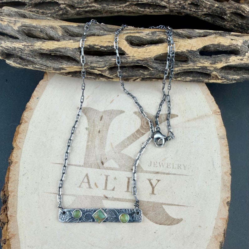 May include: A silver-toned metal necklace featuring a rectangular bar pendant with a textured surface and three small, round and square green stones. The necklace has a delicate chain link design and a lobster claw clasp closure.