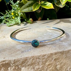 May include: A silver cuff bracelet with a round, turquoise-colored stone set in the center. The bracelet has a smooth, polished finish and is open at the ends. The bracelet is displayed on a light-colored stone surface.