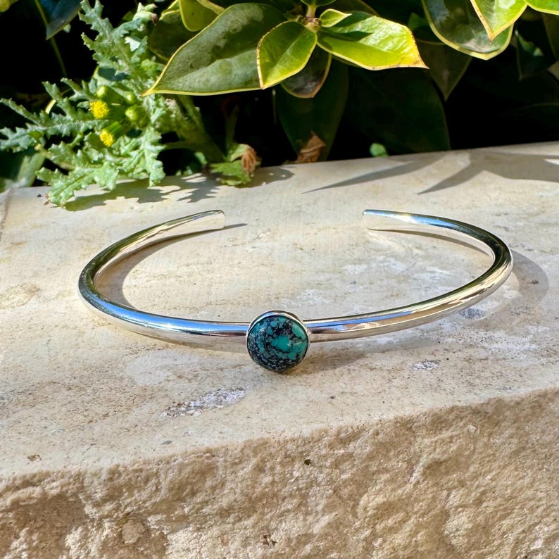 May include: A silver cuff bracelet with a round, turquoise-colored stone set in the center. The bracelet has a smooth, polished finish and is open at the ends. The bracelet is displayed on a light-colored stone surface.
