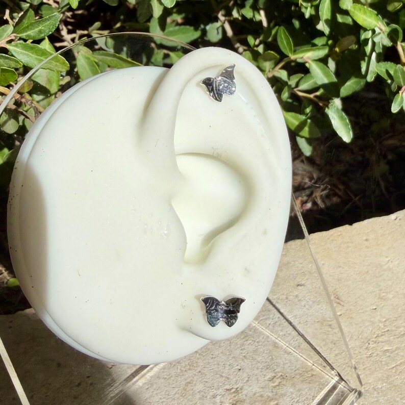 K&ouml;nnte beinhalten: Zwei silberfarbene Schmetterlings-Ohrstecker auf einem wei&szlig;en ohrf&ouml;rmigen Mannequin. Jeder Ohrring hat eine detaillierte Schmetterlingsform mit metallischem Finish und ist am oberen sowie am unteren Teil des Ohrmodells angebracht.