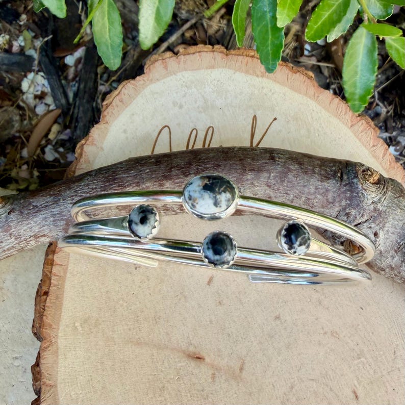 May include: Two silver-toned bangles with round, speckled, black and white stones. The bangles are stacked on a wooden surface, with a natural wood branch and green leaves in the background. The stones are set in a bezel setting.