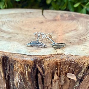 May include: A pair of silver feather cufflinks displayed on a rustic wooden surface. The cufflinks feature detailed feather designs, showcasing a delicate and natural aesthetic. The background is blurred, highlighting the cufflinks' intricate details.