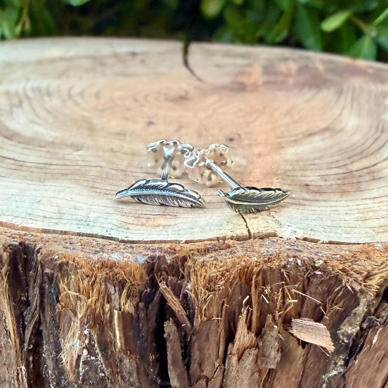 May include: A pair of silver feather cufflinks displayed on a rustic wooden surface. The cufflinks feature detailed feather designs, showcasing a delicate and natural aesthetic. The background is blurred, highlighting the cufflinks' intricate details.
