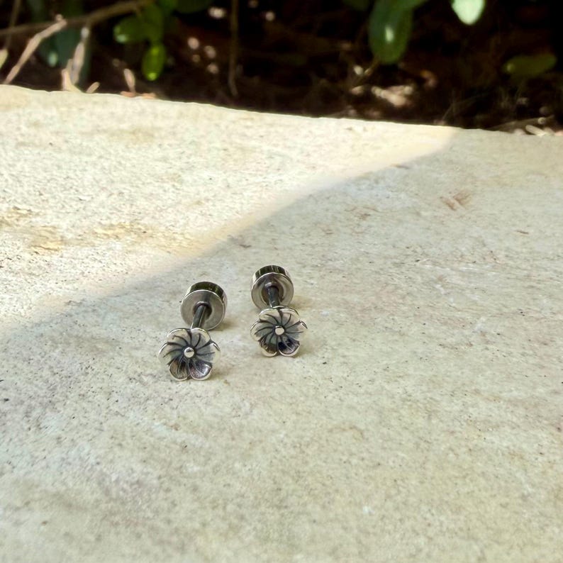 May include: A pair of silver-toned stud earrings featuring a small, six-petaled flower design. Each earring has a metallic post back and a detailed, slightly oxidized finish that highlights the individual petals of the floral shape.