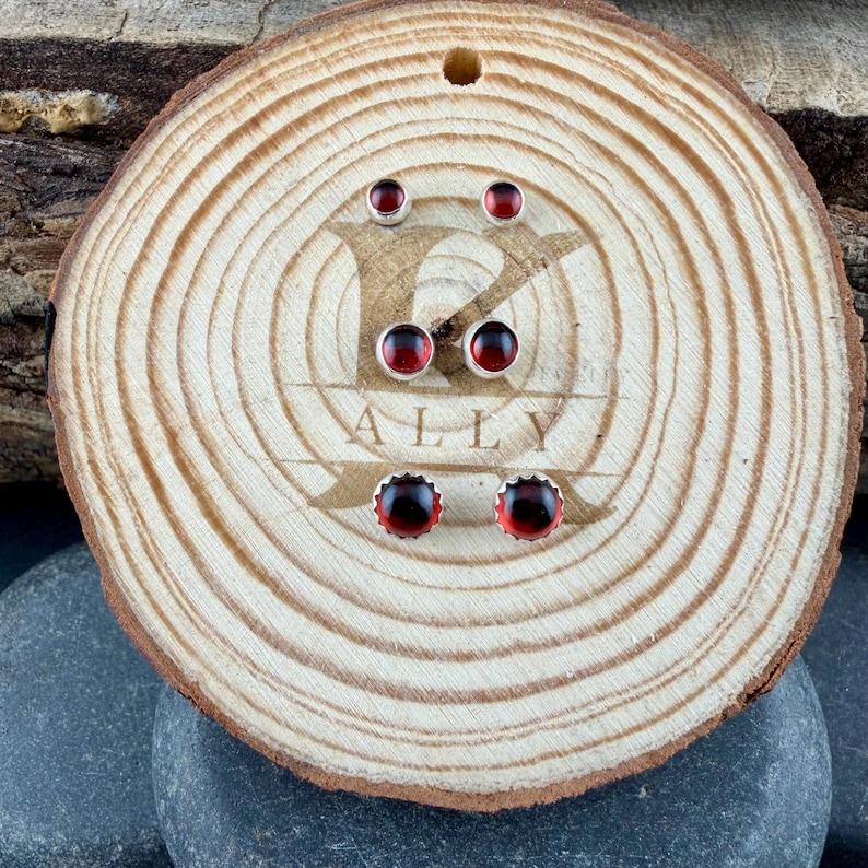 May include: Three pairs of round stud earrings featuring dark red, translucent stones set in silver-toned metal. The earrings are arranged vertically on a circular wooden slice with the engraved text K ALLY visible in the center of the wood grain.