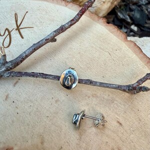 May include: Silver-coloured cowboy hat stud earrings. The earrings are shaped like a hat, with a curved brim and a central indentation. The earrings are displayed on a wooden surface with a twig.