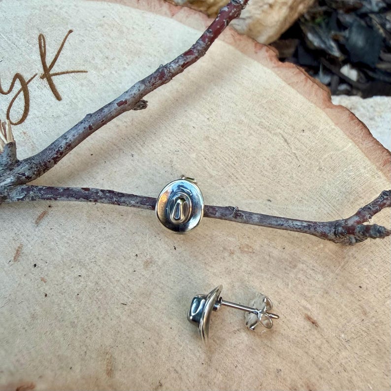 May include: Silver-coloured cowboy hat stud earrings. The earrings are shaped like a hat, with a curved brim and a central indentation. The earrings are displayed on a wooden surface with a twig.