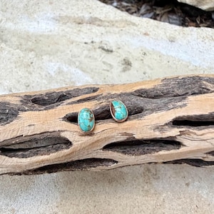 May include: A pair of turquoise stud earrings with a silver setting. The earrings have an oval shape and are set on a piece of driftwood. The turquoise has a mottled pattern of blue and brown.