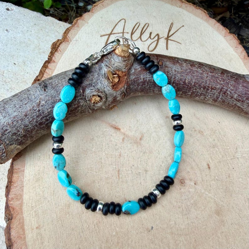 May include: A turquoise and black beaded bracelet with a silver clasp. The bracelet features oval turquoise beads interspersed with black beads and silver accents. The bracelet is displayed on a wooden surface.