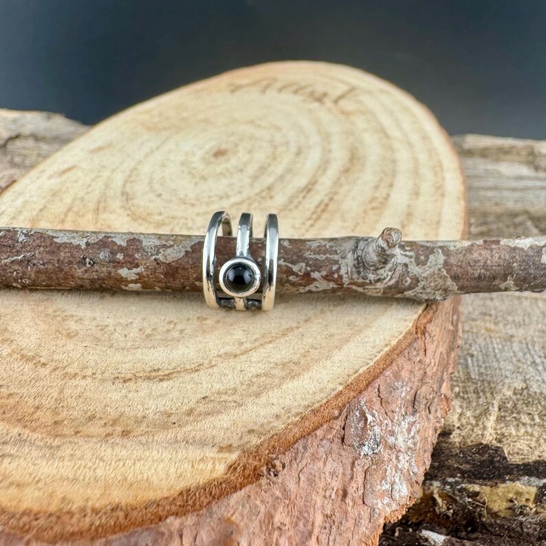 May include: A silver-colored ear cuff with a black, round stone set in the center. The cuff has a three-band design and is displayed on a small branch against a wood background.