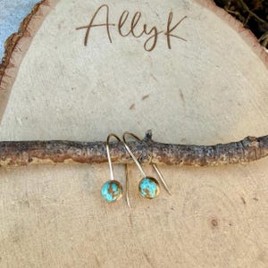 May include: A pair of gold-toned hook earrings featuring round, turquoise-colored stones with brown matrix patterns. The earrings are suspended from a thin, horizontal wooden branch resting against a light-colored wood slice engraved with the text AllyK.