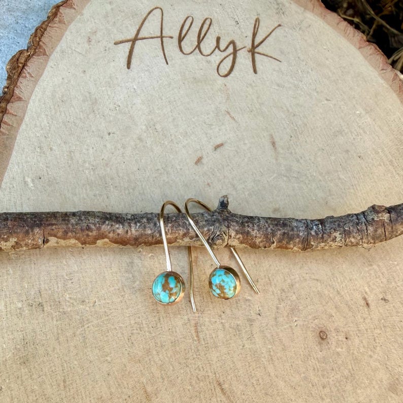 May include: A pair of gold-toned hook earrings featuring round, turquoise-colored stones with brown matrix patterns. The earrings are suspended from a thin, horizontal wooden branch resting against a light-colored wood slice engraved with the text AllyK.