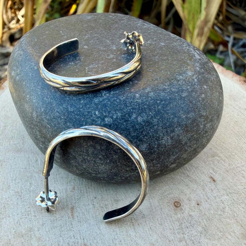 May include: Two silver hoop earrings with a textured, braided design. One earring is open, showing the post and floral-shaped closure. The other earring is closed. The earrings are displayed on a gray stone, with a wooden surface in the background.