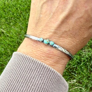 May include: A silver cuff bracelet with a hammered texture and three turquoise stones. The bracelet is worn on a wrist, showcasing the contrast between the metal and the vibrant blue-green of the stones. The bracelet is approximately 0.25 inches wide.