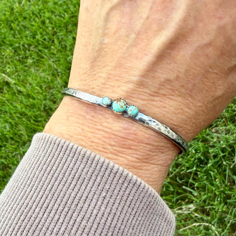 May include: A silver cuff bracelet with a hammered texture and three turquoise stones. The bracelet is worn on a wrist, showcasing the contrast between the metal and the vibrant blue-green of the stones. The bracelet is approximately 0.25 inches wide.