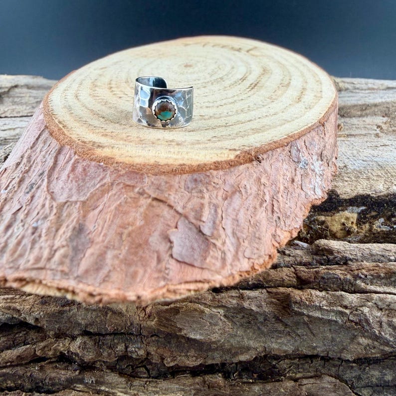 May include: A silver-toned ring with a hammered texture and a turquoise-colored stone. The ring is displayed on a wooden surface, showcasing its handcrafted details and unique design. The ring is a piece of jewelry.