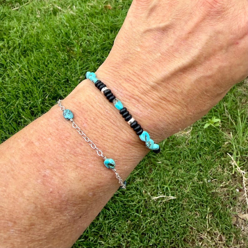 May include: Two bracelets are displayed on a wrist against a backdrop of green grass. One bracelet is silver with turquoise beads. The other bracelet has black beads, silver accents, and turquoise beads.