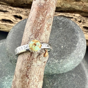 May include: A silver ring with a round, light turquoise stone set in a scalloped bezel. The band features etched designs. The ring is displayed on a wooden branch against a blurred background of rocks.