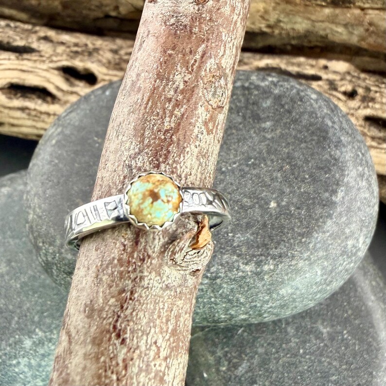May include: A silver ring with a round, light turquoise stone set in a scalloped bezel. The band features etched designs. The ring is displayed on a wooden branch against a blurred background of rocks.