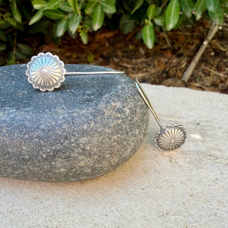May include: A pair of silver-toned earrings featuring a circular, sunburst-inspired design with radiating lines and a scalloped edge. Each earring is attached to a thin, straight metal post, showcasing a metallic finish with subtle dark oxidation.