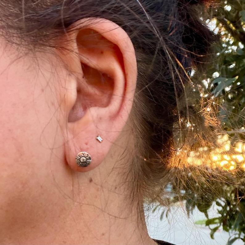 May include: A silver-toned stud earring shaped like a sun with radiating rays sits in the earlobe, accompanied by a tiny, rectangular-shaped silver-toned stud earring positioned slightly higher up on the ear cartilage.