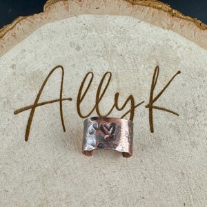 May include: A copper-colored ring with a hammered texture and a heart design. The ring is displayed on a wooden surface with the handwritten text "Allyk". The ring has a rustic, handmade appearance.
