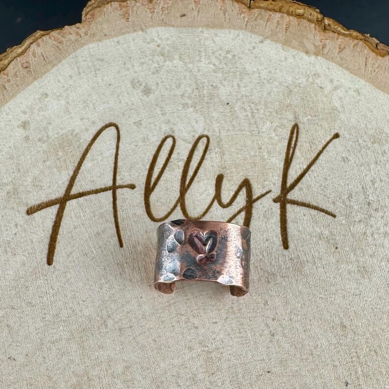 May include: A copper-colored ring with a hammered texture and a heart design. The ring is displayed on a wooden surface with the handwritten text "Allyk". The ring has a rustic, handmade appearance.