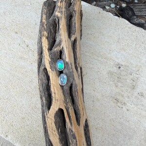 May include: Two oval-shaped gemstone earrings with silver settings. One earring has a vibrant green and blue color, while the other has a soft blue hue. The earrings are displayed on a weathered, textured piece of wood.