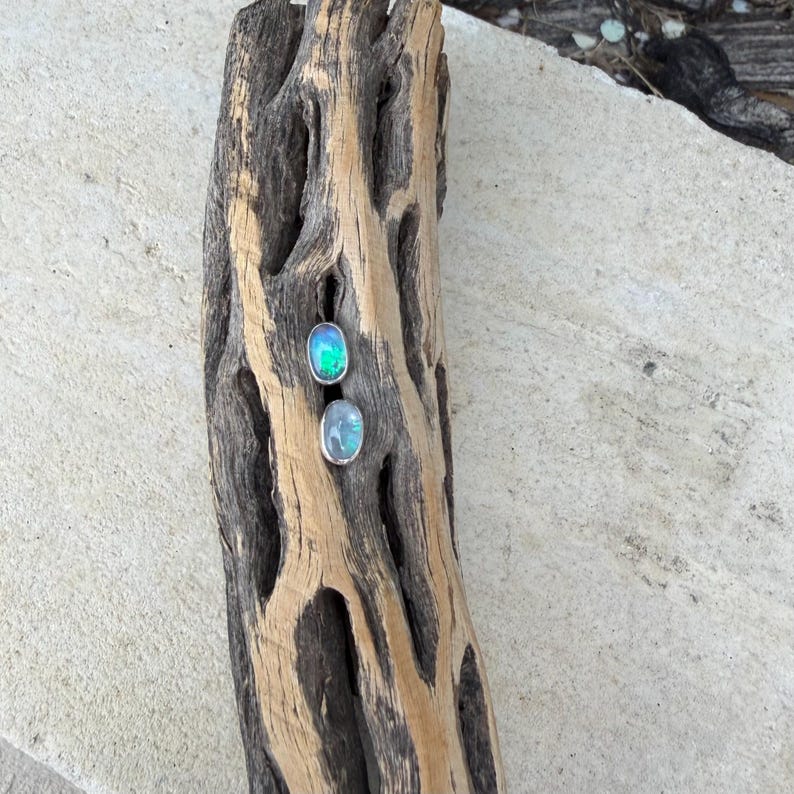 May include: Two oval-shaped gemstone earrings with silver settings. One earring has a vibrant green and blue color, while the other has a soft blue hue. The earrings are displayed on a weathered, textured piece of wood.