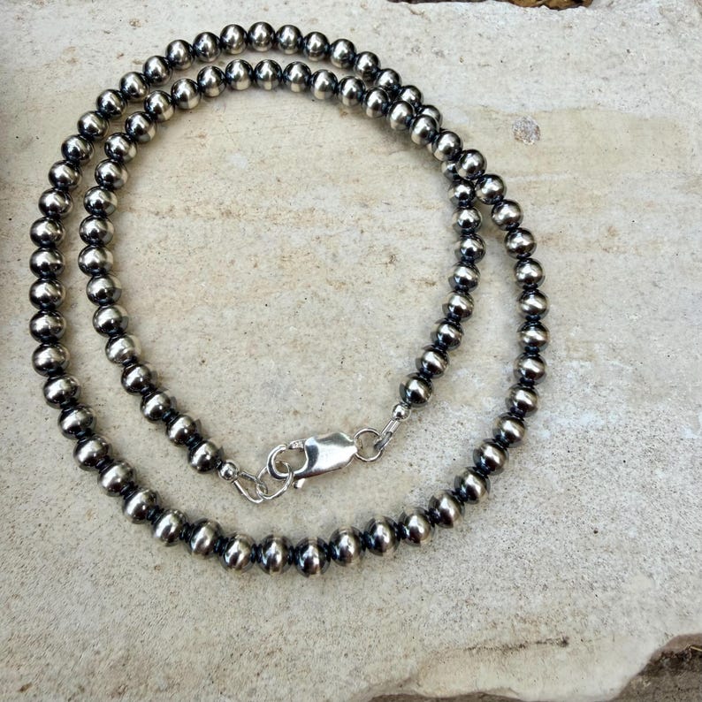 Pu&ograve; includere: Una collana di perline d'argento con chiusura a moschettone. La collana &egrave; composta da piccole perline rotonde d'argento infilate insieme. Le perline hanno una finitura metallica lucida. La collana &egrave; esposta su una superficie chiara e strutturata.