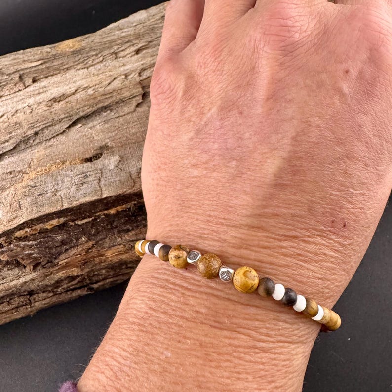 May include: Beaded bracelet featuring a mix of brown, white, and silver beads. The bracelet has a natural, earthy aesthetic, with varying sizes and textures of beads. The bracelet is worn on a wrist.