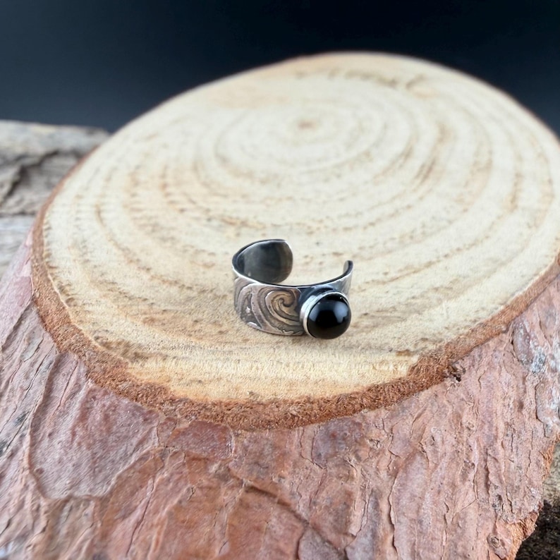 May include: A silver-toned adjustable ring with a black gemstone. The ring features an engraved wave pattern on the band. The black gemstone is round and set in a bezel setting. The ring is displayed on a wooden surface.