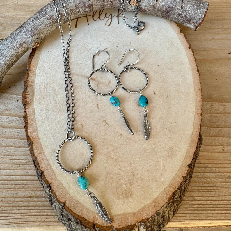 May include: A silver necklace and earrings set with turquoise stones and feather charms. The necklace features a large circular pendant with a turquoise stone and a dangling feather charm. The earrings have a similar design, with turquoise stones and feather charms.