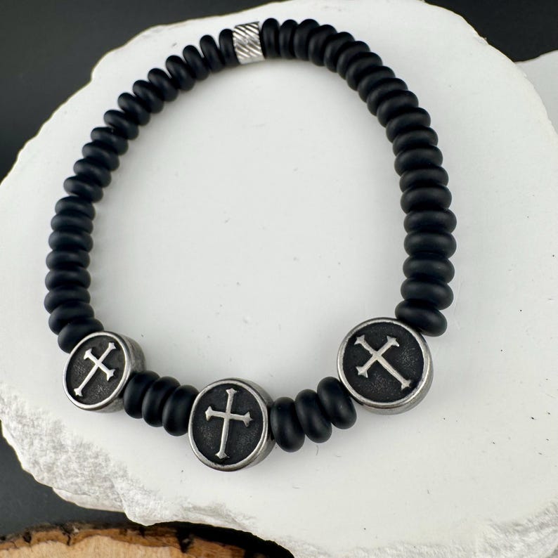 May include: Black beaded bracelet featuring three silver-toned cross charms. The bracelet has a silver accent bead. The beads are a matte black color, and the crosses have a detailed design.