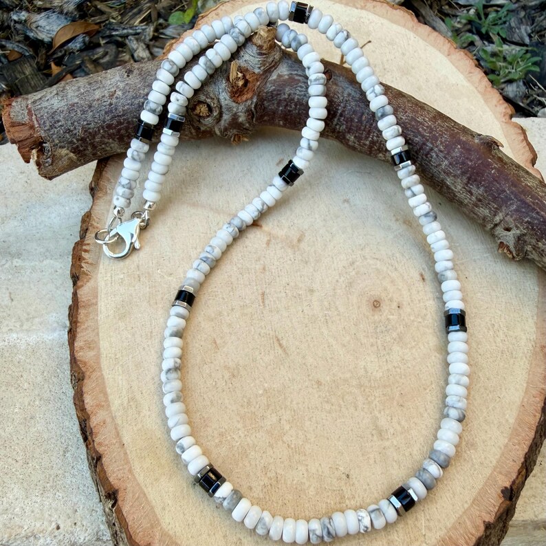 May include: White and gray beaded necklace with black accents. The necklace features small, round beads in shades of white and gray, interspersed with black cylindrical beads. A silver-colored clasp secures the necklace.