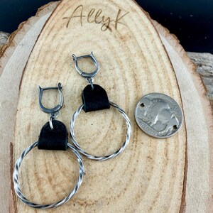 May include: Silver hoop earrings with a twisted design and black leather accents. A silver coin with a buffalo image is next to the earrings. The earrings are displayed on a wooden surface. The name "Allyk" is written on the wood.