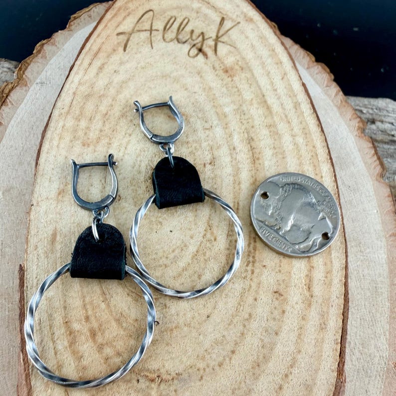 May include: Silver hoop earrings with a twisted design and black leather accents. A silver coin with a buffalo image is next to the earrings. The earrings are displayed on a wooden surface. The name "Allyk" is written on the wood.