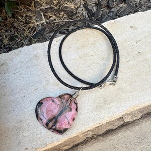 May include: A heart-shaped pendant necklace featuring a pink and black stone. The stone is wire-wrapped and suspended from a black braided cord necklace with a silver-tone clasp. The pendant is approximately 2 inches wide.