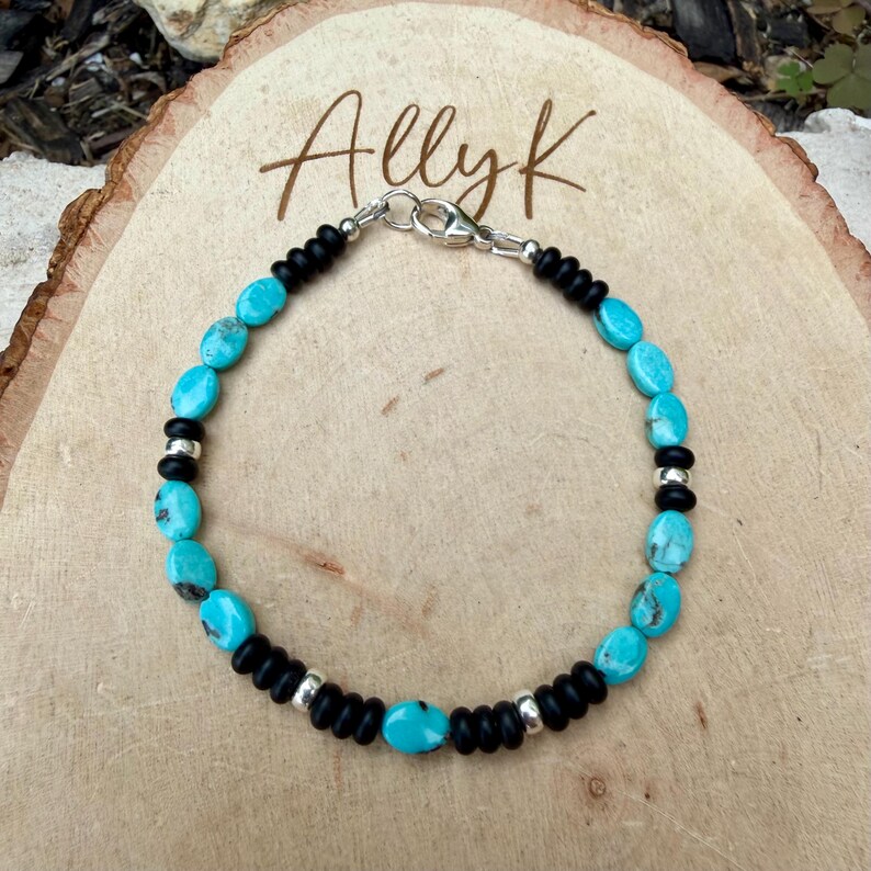 May include: A turquoise and black beaded bracelet with a silver clasp. The bracelet features oval turquoise stones interspersed with black beads and silver accents. The bracelet is displayed on a wooden surface, with the name "Allyk" written in cursive.