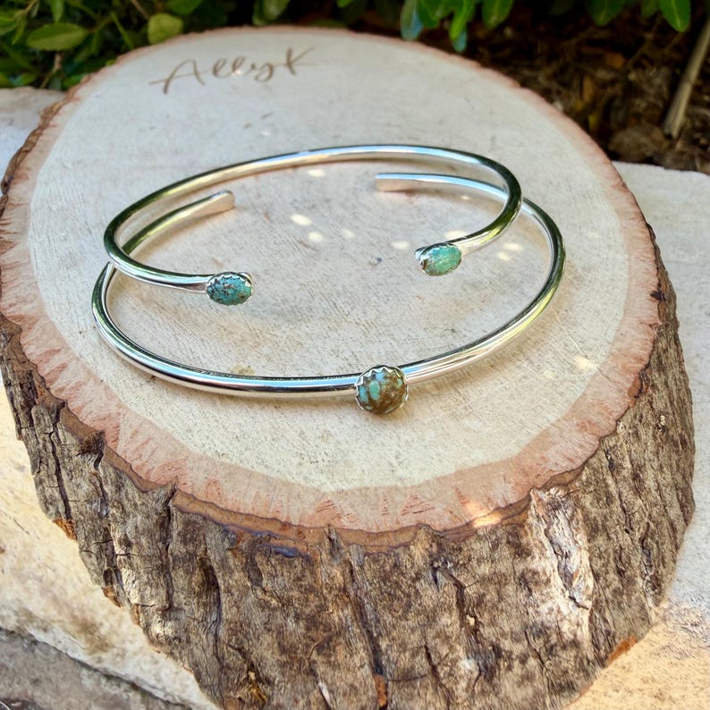 May include: Two silver-toned cuff bracelets with turquoise-colored stones. The bracelets are displayed on a natural wood slice, showcasing the jewelry's design and texture. The bracelets are open-ended, allowing for easy wear.