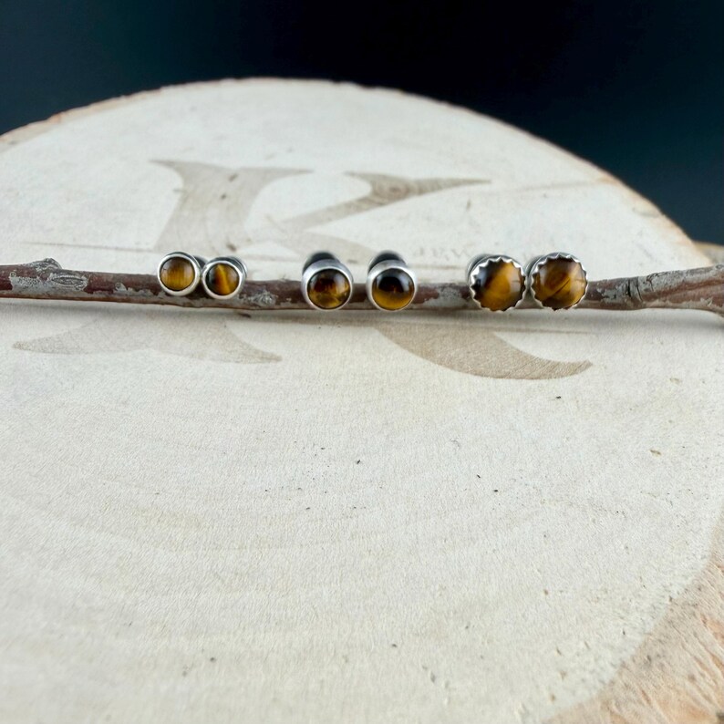 Puede incluir: Tres pares de pendientes de bot&oacute;n redondos con piedras preciosas de ojo de tigre pulidas engastadas en metal plateado. Cada pendiente muestra las caracter&iacute;sticas bandas doradas y marrones, dispuestas en fila sobre una rama de madera.
