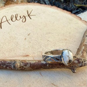 May include: A silver ring with an oval, white and brown stone. The ring band has a decorative pattern. The ring is displayed on a small branch, with the name "Allyk" written on a wooden surface in the background.