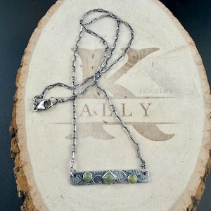 May include: A silver-toned necklace featuring a rectangular bar pendant with an oxidized finish, textured patterns, and three inlaid green stones. The center stone is square-shaped, flanked by two round stones. The chain is a linked cable style.